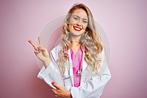 Young beautiful doctor woman using stethoscope over pink isolated background smiling with happy face winking at the camera doing