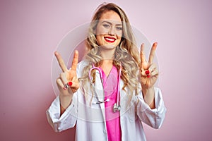 Young beautiful doctor woman using stethoscope over pink isolated background smiling looking to the camera showing fingers doing