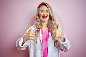 Young beautiful doctor woman using stethoscope over pink isolated background success sign doing positive gesture with hand, thumbs