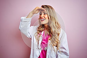 Young beautiful doctor woman using stethoscope over pink isolated background very happy and smiling looking far away with hand