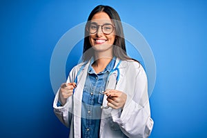 Young beautiful doctor woman wearing stethoscope and glasses over blue background pointing fingers to camera with happy and funny