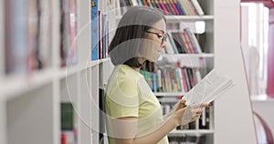 Young beautiful european female student wearing glasses is reading book in college library. Side view.