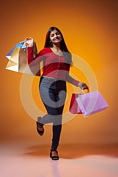 Young beautiful excited girl, student with bright shopping packages isolated on yellow studio background in neon light
