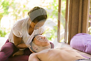 Young beautiful and exotic Asian Thai therapist woman giving traditional head and facial Balinese massage to Caucasian man at