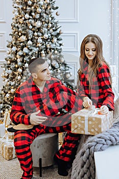 A young beautiful family couple in the same red pajamas, near the Christmas tree, rejoice and exchange New Year's gifts.