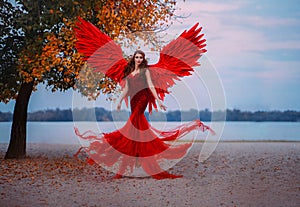 Young beautiful fantasy woman fallen angel lying in air near a tree with orange leaves. Creative red costume, huge