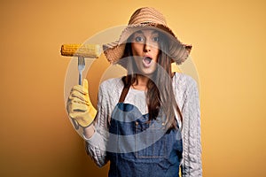 Young beautiful farmer woman wearing apron and hat holding fork with cob corn scared in shock with a surprise face, afraid and