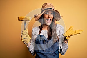 Young beautiful farmer woman wearing apron and hat holding fork with cob corn very happy and excited, winner expression