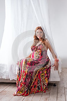 Young beautiful fashion pregnant woman in colorful dress sitting on a bed