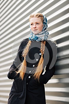 Young beautiful fashionable redhead woman with braids hairdo in blue white headcraft stylish denim black trench jacket posing outd