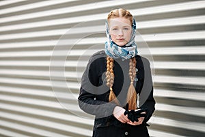 Young beautiful fashionable redhead woman with braids hairdo in blue white headcraft stylish denim black trench jacket posing outd