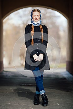 Young beautiful fashionable redhead woman with braids hairdo in blue white headcraft stylish denim black trench jacket posing outd