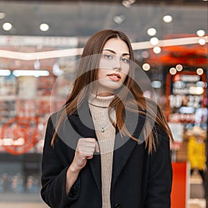 Young beautiful fashionable woman brunette in trendy beige shirt in black elegant coat posing in the modern mall. Attractive