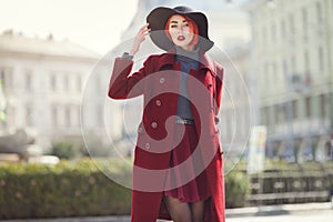 Young beautiful fashionable woman posing on the street. Model wearing stylish black wide-brimmed hat, red coat. Girl