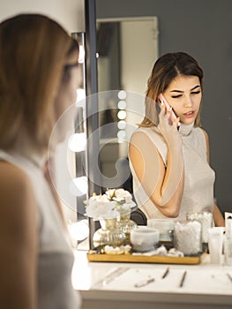 Young beautiful female actress with her reflection in the make-up mirror in the dressing room talking on the cell phone