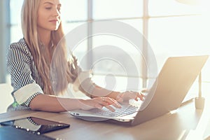 Young beautiful female copywriter typing texts and blogs in spacious light office, her workplace, using pc keyboard