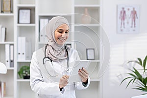 Young beautiful female doctor in hijab using tablet computer, doctor worker in white medical coat and stethoscope inside
