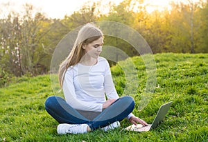 Young beautiful female student sitting on grass and typing on a laptop  in spring