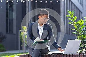 Young beautiful female student studying, remotely in the park, sitting on a bench, online and remotely, using laptop, near college