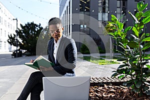 Young beautiful female student studying, remotely in the park, sitting on a bench, online and remotely, using laptop, near college