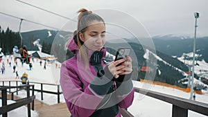 Young beautiful female tourist uses a mobile phone on the lift of a ski resort, mountains and skiers in the background.