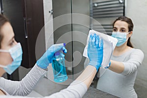 Young beautiful female wearing protective face mask and cleaning mirror in bathroom. Coronavirus protection prevention