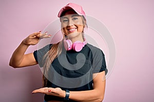 Young beautiful fitness sports woman wearing training cap and headphones over pink background gesturing with hands showing big and