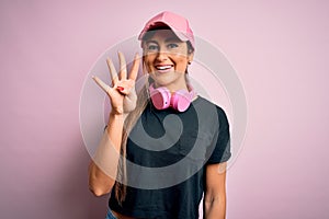 Young beautiful fitness sports woman wearing training cap and headphones over pink background showing and pointing up with fingers
