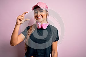 Young beautiful fitness sports woman wearing training cap and headphones over pink background smiling and confident gesturing with