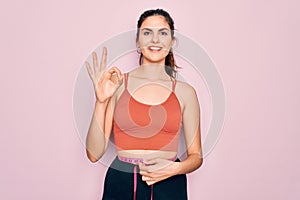 Young beautiful fitness woman wearing sport excersie clothes using measuring tape doing ok sign with fingers, excellent symbol