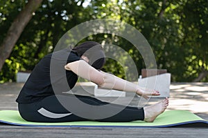 Young beautiful flexible girl makes a lean forward with her hands from a sitting position. Yoga class in the morning in the park.