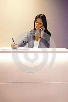 Young beautiful focused receptionist speaks on phone and writes down notes about client
