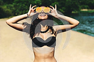 Young beautiful funny summer model holding an orange in front of her eyes smiles and laughs on the beach in a bikini on a sunny su