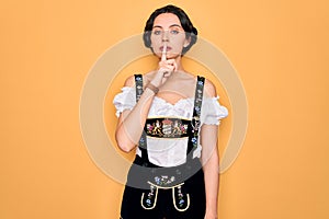 Young beautiful german woman with blue eyes wearing traditional octoberfest dress asking to be quiet with finger on lips