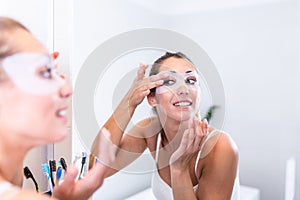 Young beautiful girl applying facial mask on skin. Looking in the mirror in bathroom, Wrapped in a towel, having fun