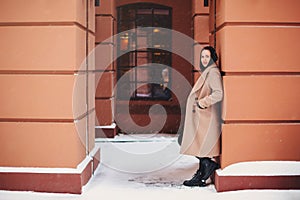 Young beautiful girl in beige coat and leather boots lonely standing outside in snowy winter weather