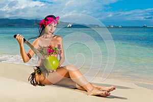 Young beautiful girl in a bikini made of flowers sitting on a tropical beach and coconut with a machete. Blue sea in the backgroun
