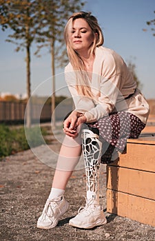 A young beautiful girl with a bionic prosthesis sits on a bench in the park.