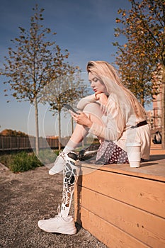 A young beautiful girl with a bionic prosthesis sits in the park and looks at a smartphone.