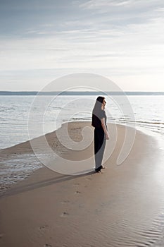 Young beautiful girl in black clothes walking on the beach. She walks along shore. A walk at sunset. Nobody around