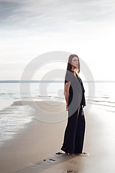 Young beautiful girl in black clothes walking on the beach. She walks along shore. A walk at sunset. Nobody around