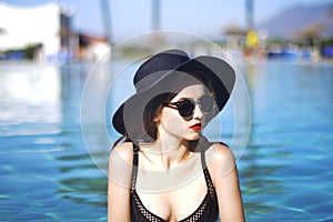 Young beautiful girl in black fashion hat, velvet skin, red lips, black swimsuit posing in the pool in blue water, stylish sungla