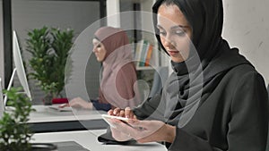 Young beautiful girl in black hijab sits in office and uses smartphone. Girl in black hijab in the background. Arab