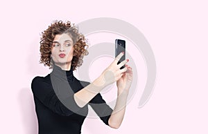 Young beautiful girl in a black turtleneck makes a selfie with a smartphone pouting her lips, on light pink background