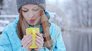 Young beautiful girl blowing on a hot tea or coffee standing on the shore of a winter lake.