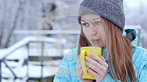 Young beautiful girl blowing on a hot tea or coffee standing on the shore of a winter lake.