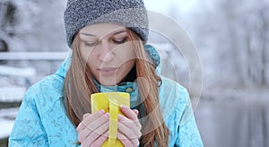 Young beautiful girl blowing on a hot tea or coffee standing on the shore of a winter lake.