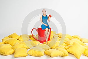 Young beautiful girl in blue dress sits on a huge cup in a yellow pillows and holds cup of coffee on a white background