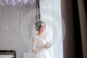 Young beautiful girl bride in a peignoir near the window sill with a cup of coffee and is waiting for the groom, morning of the