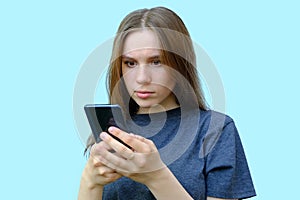 young beautiful girl with brown eyes isolated on a blue background. The teenager looks at the phone with a surprised